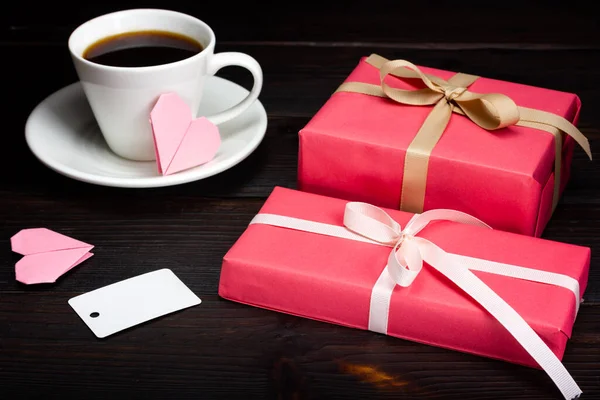 Deux cadeaux enveloppés dans du papier cadeau rose, une tasse de café et des étiquettes en papier vierge sur une table en bois sombre. — Photo