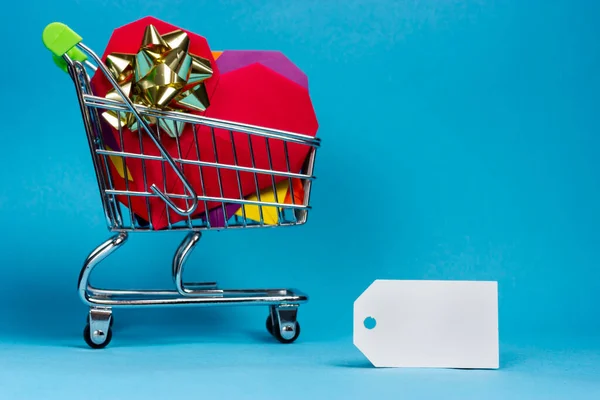 Carrinho cheio de presentes e etiqueta de papel em branco em um fundo azul claro. Comprar presentes. Lugar para texto. — Fotografia de Stock