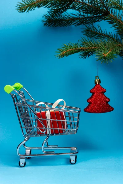 Um carrinho com presentes e um brinquedo de árvore de Natal em um ramo de abeto. Comprar presentes. — Fotografia de Stock