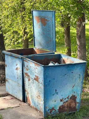 Parktaki paslı mavi çöp konteynırında ağaçlar ve çimenler var. Yaratıcı ve ticari kullanım için ideal