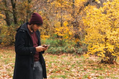 Parkta duran beyaz bir adam, sonbaharda cep telefonundan mesaj yolluyor.