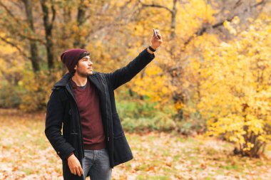 Tanınan bir adam doğada fotoğraf çekiyor. Sonbaharda kalın giysilerle ayakta duruyor.