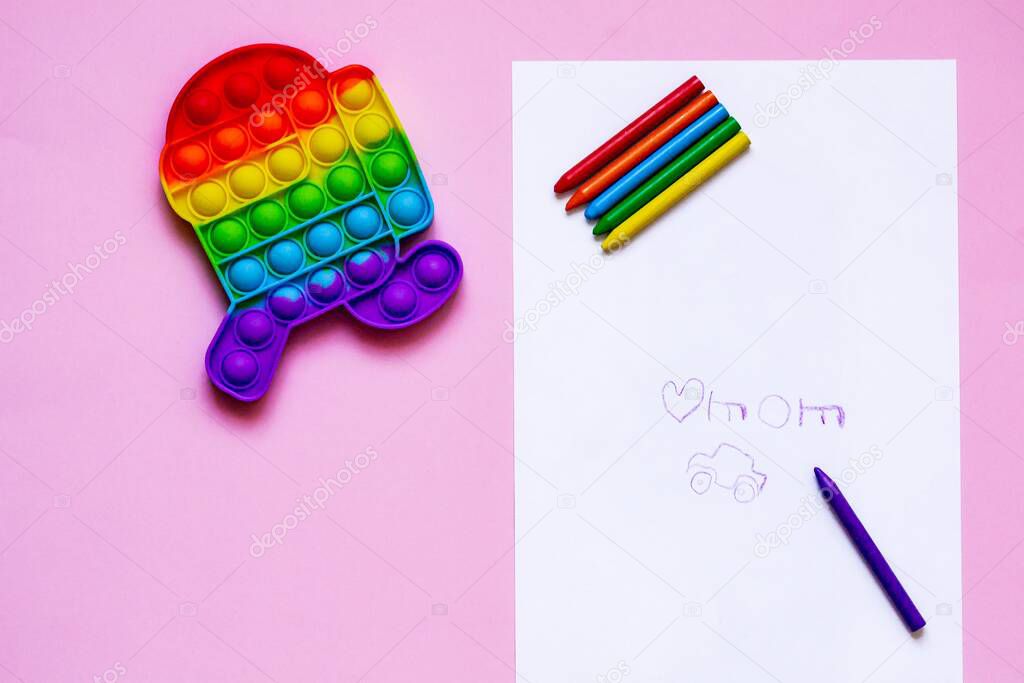 Psychological health concept. Anti-stress sensory color push fidget toy to develop fine motor skills. Flat lay on pink background with colored pencils and childish drawing.