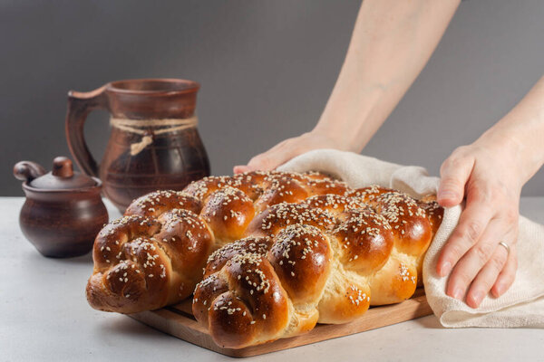 Woman holding just baked Challah bread with poppy and sesame seeds. Covered with white cover. On white and grey background.