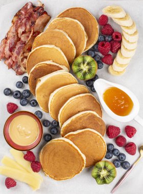 Kahvaltı panosu: krep, pastırma, peynir, fıstık ezmesi, bal, taze meyve: ahududu, yabanmersini, kivi, muz. Açık gri arkaplan. Düz yatıyordu.