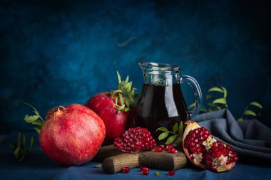 Pomegranate juice in a jug, whole pomegranates and seeds on dark blue background. Dark and moody. clipart