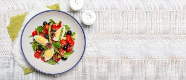 Ançüezli Fransız Nicoise salatası, yeşil fasulye, domates, yumurta, siyah zeytin, kapari, beyaz tabak ve beyaz ahşap arka plan. Üst görünüm, pankart boyutu. 