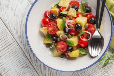 Vejetaryen Akdeniz salatası. Haşlanmış patates, domates, soğan, kekik, kapari, siyah zeytin, fesleğen yaprağı. Koyu ahşap arkaplan, üst manzara.