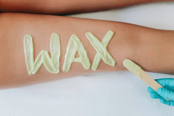 Top view on inscription wax laid out with hot wax for depilation. The gloved hand of the depilation master holding spatula with wax. Hair removal procedure on female thighs. The concept of epilation