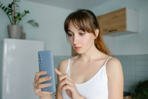 Young pretty girl with the bangs reading a message or shopping online in a smartphone at home in blue interior