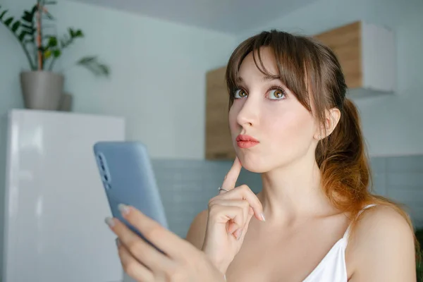 Portrait of a young pensive girl with the bangs holding smartphone and looking away with hand on her chin at home in blue interior.