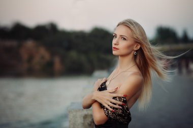Beautiful young fashionable woman posing in dress at the river coast in the evening after sunset. Vogue style. Urban background