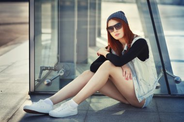 Outdoor lifestyle fashion portrait of pretty young sitting girl, wearing in hipster swag grunge style urban background. Red hair. Retro vintage toned image, film simulation.