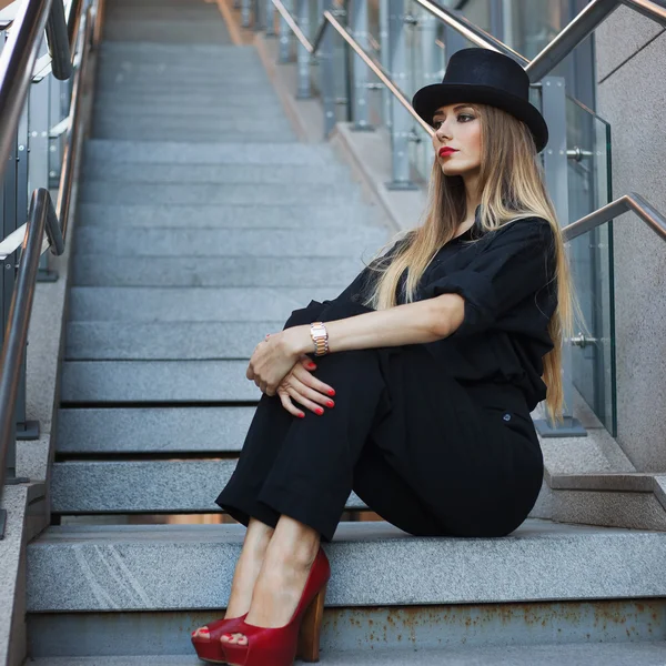 Belle jeune femme à la mode posant en suite noire, chaussures rouges à talons hauts et chapeau noir. Style vogue. Contexte urbain — Photo