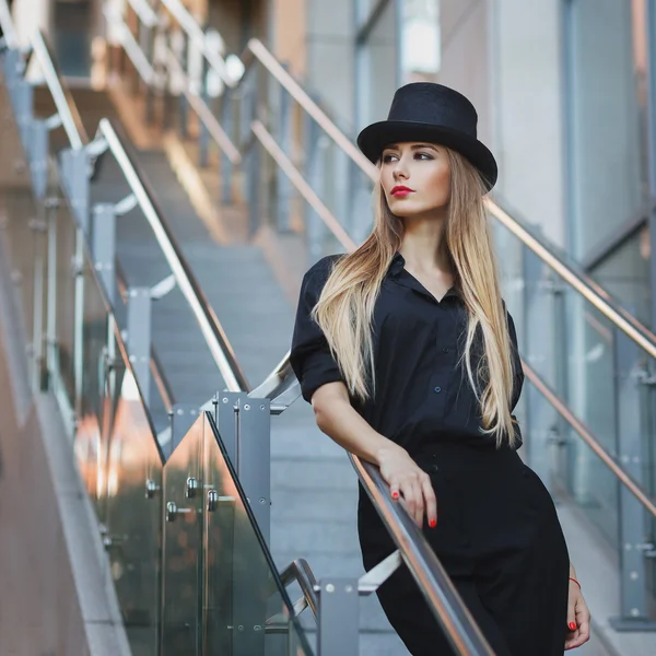Schöne junge modische Frau posiert in schwarzen Anzügen, roten Schuhen mit hohen Absätzen und schwarzem Hut. modischen Stil. Urbaner Hintergrund — Stockfoto