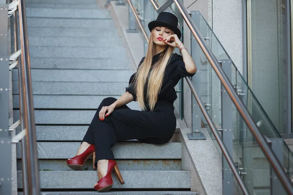 Όμορφη νέα μοντέρνα γυναίκα που ποζάρει στη μαύρη σουίτα, κόκκινα παπούτσια με ψηλά τακούνια και μαύρο καπέλο. Στυλ μόδας. Αστικό υπόβαθρο — Φωτογραφία Αρχείου
