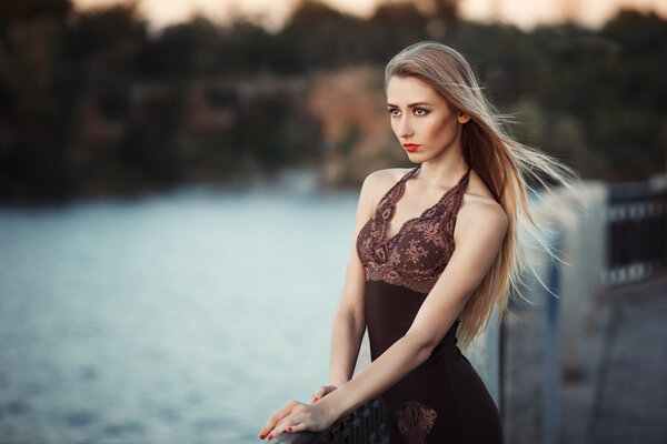 Beautiful young fashionable woman posing in dress at the river coast in the evening after sunset. Vogue style. Urban background