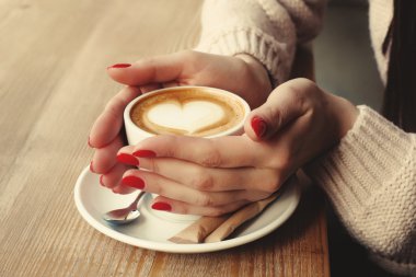 Fincan kahve kapuçino desen kalp ile köpük ile tutarak yakın çekim eller. Mükemmel kırmızı jel Lehçe manikür. Ahşap doğal masa. Yaratıcı renk sıcak mesaj işleme instagram tarzı.