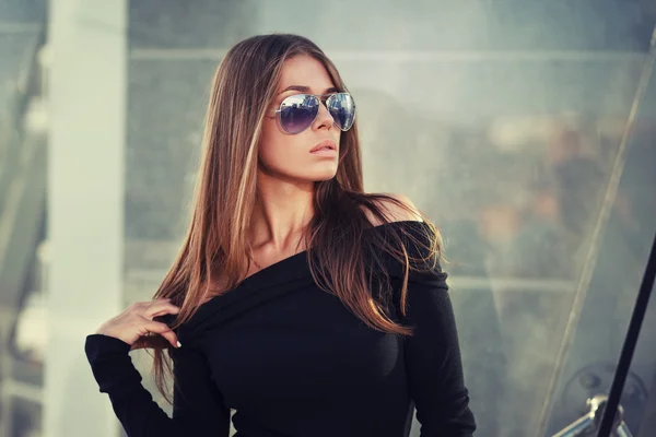 Hermosa joven morena en bonito vestido negro, gafas de sol. Posando sobre fondo urbano. Foto de moda —  Fotos de Stock