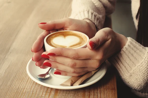 Fincan kahve kapuçino desen kalp ile köpük ile tutarak yakın çekim eller. Mükemmel kırmızı jel Lehçe manikür. Ahşap doğal masa. Yaratıcı renk sıcak mesaj işleme instagram tarzı.