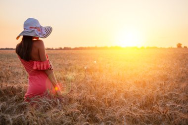 Güzel genç kadın kahverengi ile duymak gül elbise ve açık havada günbatımı üzerinde mükemmel buğday alan üzerinde güneşin isteyen zevk şapka giyiyor
