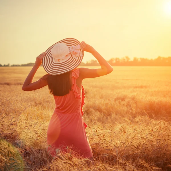 Beautiful young woman with brown hear wearing rose dress and hat with raised arms enjoying outdoors looking to the sun on perfect wheat field on sunset