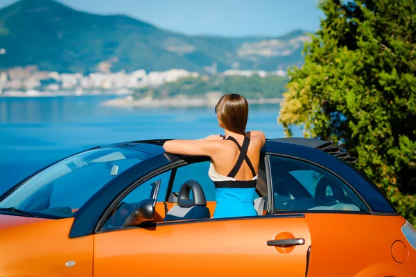 Belle jeune femme aux cheveux longs debout à l'arrière avec du cabriolet orange sur la côte méditerranéenne — Photo
