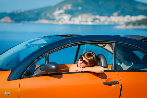 Schöne junge Frau mit langen Haaren sitzt und lächelt im orangefarbenen Cabrio an der Mittelmeerküste — Stockfoto