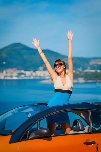 Beautiful young woman with long hair standing in orange cabriolet at the Mediterranean sea coast — Stock Photo, Image