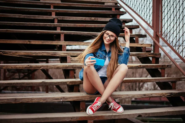 Ung sexig blondin hipster kvinna poserar för selfie och skrattar. Klädd i jeansjacka, hipster svart hatt och glasögon. Livsstil porträtt — Stockfoto
