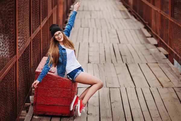 Jeune femme hipster sexy assise sur une boîte vintage rouge et riant. Veste en jean, chapeau noir hipster et lunettes. Mode de vie ville portrait.a — Photo