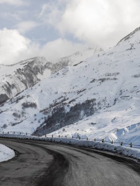 Etrafı karla kaplı engin tepelerle çevrili asfalt dağ yolu bulutlu bir kış gökyüzü altında sakin ve görkemli bir alp manzarasını çağrıştırıyor..