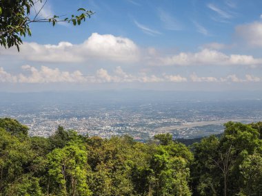 Tayland, Chiang Mai 'nin panoramik manzarası, dağ açısından. Ağaçların yemyeşil tepeleri, bulutlu ve aydınlık bir günde sisli ufka uzanan genişleyen şehir manzarasını çerçeveler. Bir havaalanı pisti görünür
