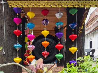 Çeşitli şekil ve kalıplarda canlı ipek fenerler geleneksel ahşap bir zemin üzerinde asılı, büyük bir gong ve arka planda görünen Tayland tapınağı mimarisi, zengin sergiler