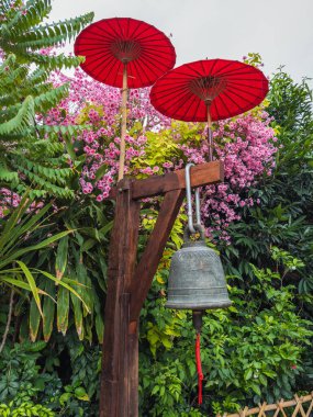 Geleneksel bir Tayland bronz çanı ve iki parlak kırmızı şemsiyenin canlı bir sahnesi. Berrak tropikal yapraklar ve sakin bir bahçede pembe çiçekler arasında..