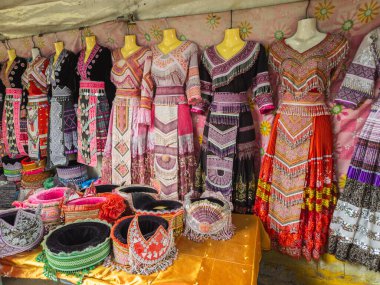 Canlı geleneksel etnik elbiseler ve mankenler ve bir market tezgahında sergilenen karmaşık başlıklar. Zengince işlenmiş kumaşlar kültürel mirası ve zanaatkarlığı sergiliyor..