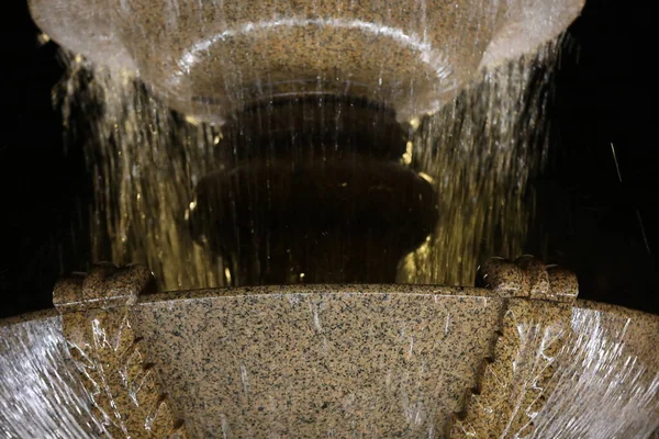 Night water fountain.Decorative stone vase with waterfall close up.Blurred streams of water from the upper Cup overflow into the lower part.Grey granite with black flecks wet glossy.Background image