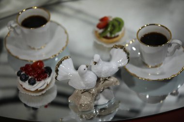 İki fincan kahvenin bulanık zemininde bir çift porselen beyaz güvercin ve aynalı bir masada üzerinde bir yansıma olan renkli pastalar. Sevginin ve mutluluğun sembolü.