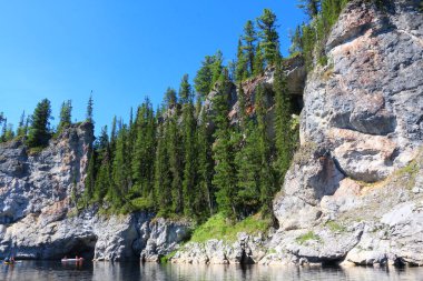 Sudan kayalık kıyılara mavi gökyüzünün altında uzun çam ağaçları ve mağaranın girişinde kayık kanoları olan dağın tabanında bir delik. Vahşi doğanın manzarası.
