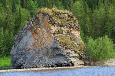 Suyun yakınındaki çakıl taşı plajının kıyısındaki ağaçlar arasında tek bir taş bloğun görüntüsü. Çok renkli, pürüzsüz duvarları olan turuncu gri katmanlı kaya. Kayanın fotoğrafı.