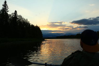 Kafasının ve gövdesinin silueti, alacakaranlıkta gölde kürek çeken mavi gökyüzünde ışıldayan bulutlar ve altın rengi dalgalarla gün batımını seyreden bir adama ait. İnsan güzelliğe bakar.
