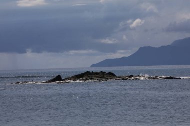 Deniz, taştan bir adada kuşla parıldayan su ve ufukta belirsizlik olan dağlar. Güzel deniz manzarası