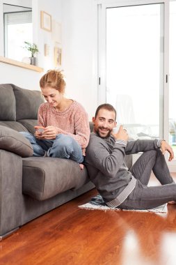 Mutlu, genç bir çift kahve fincanları tutarak rahatlıyor ve oturma odasında koltukta birlikte telefon kullanıyorlar..