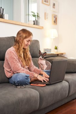 Dizüstü bilgisayarda yazan kadın, kedisiyle kanepede oturan..