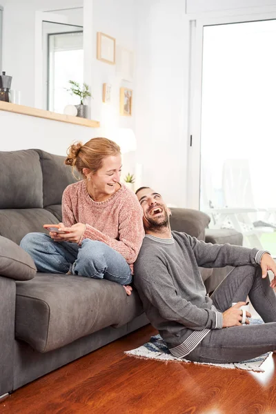 Mutlu genç çift ellerinde kahve fincanları tutarak gülüyor ve oturma odasında birlikte kanepede telefon kullanıyorlar..