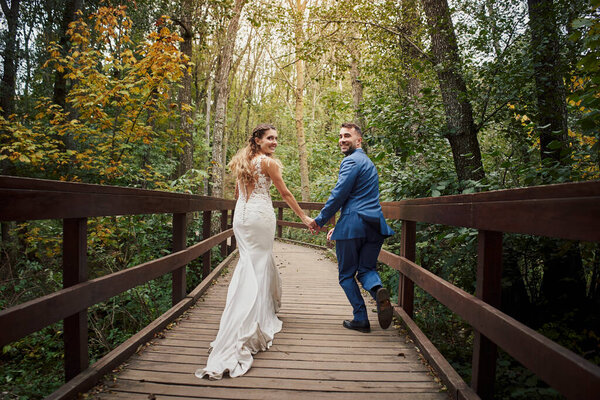 Back view of a newly married couple running and looking back over a bridge in the woods.