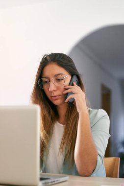 Evde dizüstü bilgisayarla çalışan ve telefonda konuşan bir kadının ön görüntüsü. Dikey fotoğraf.