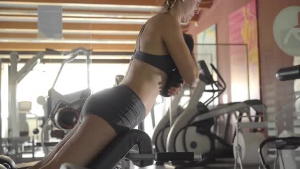 スポーツシミュレーターでジムでフィットネスに従事する美しい若い女性が背中とお尻の筋肉を振る — ストック動画