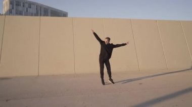 Siyah giyinmiş atletik genç adam, şehir dışında egzersiz yaparken gri bir duvarın önünde takla atıyor..