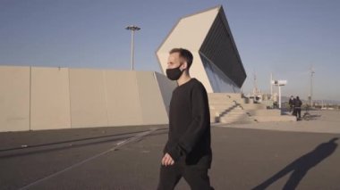 Siyah elbiseli ve yüz maskeli atletik genç adam takla atıyor ve sonra şehir dışında park çalışması yaparken koşuyor..